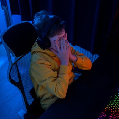 Ein Junge sitzt in einem dunklen Zimmer an einem Schreibtisch und spielt Online-Games, er hält erschrocken die Hände vors Gesicht.