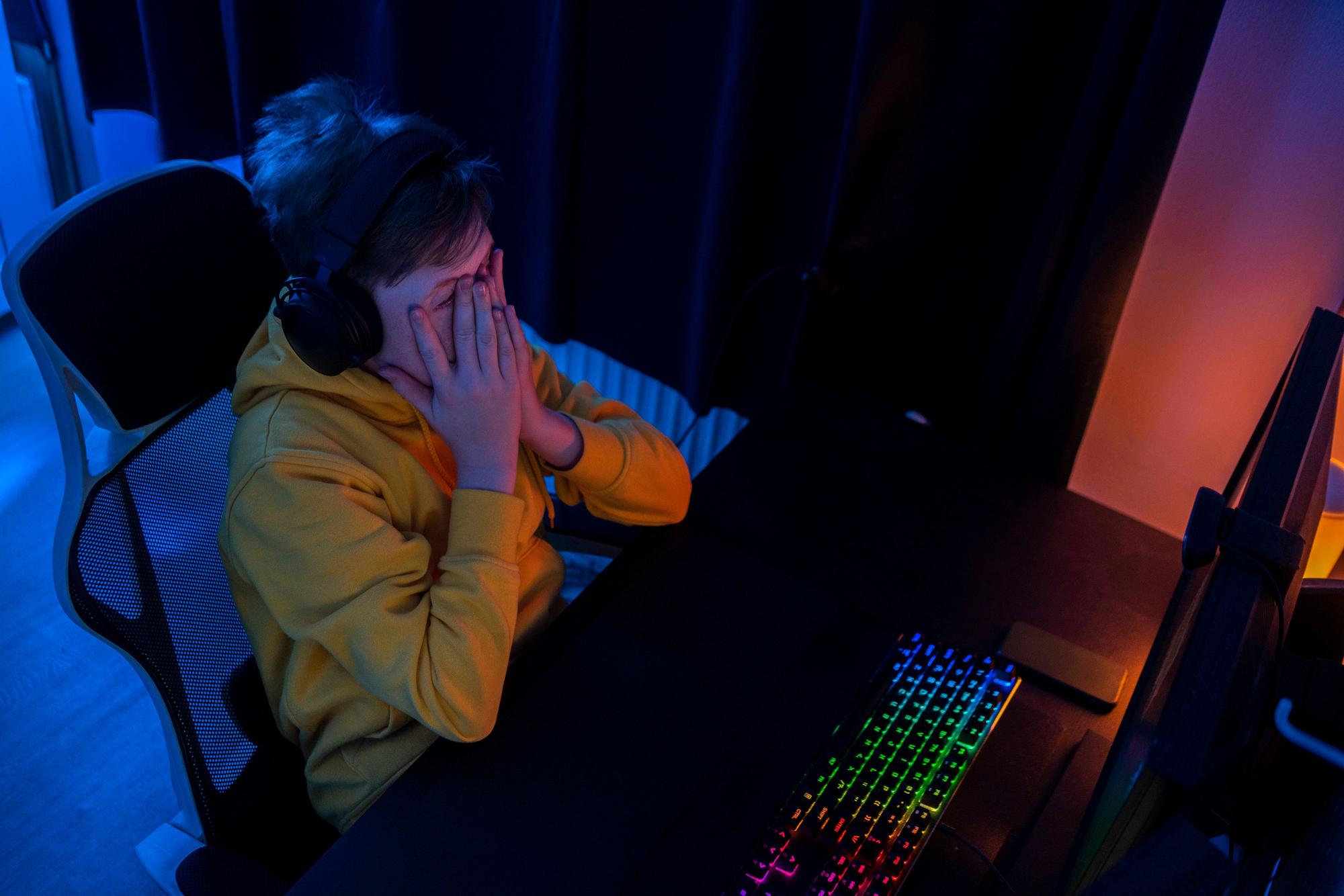 Ein Junge sitzt in einem dunklen Zimmer an einem Schreibtisch und spielt Online-Games, er hält erschrocken die Hände vors Gesicht.