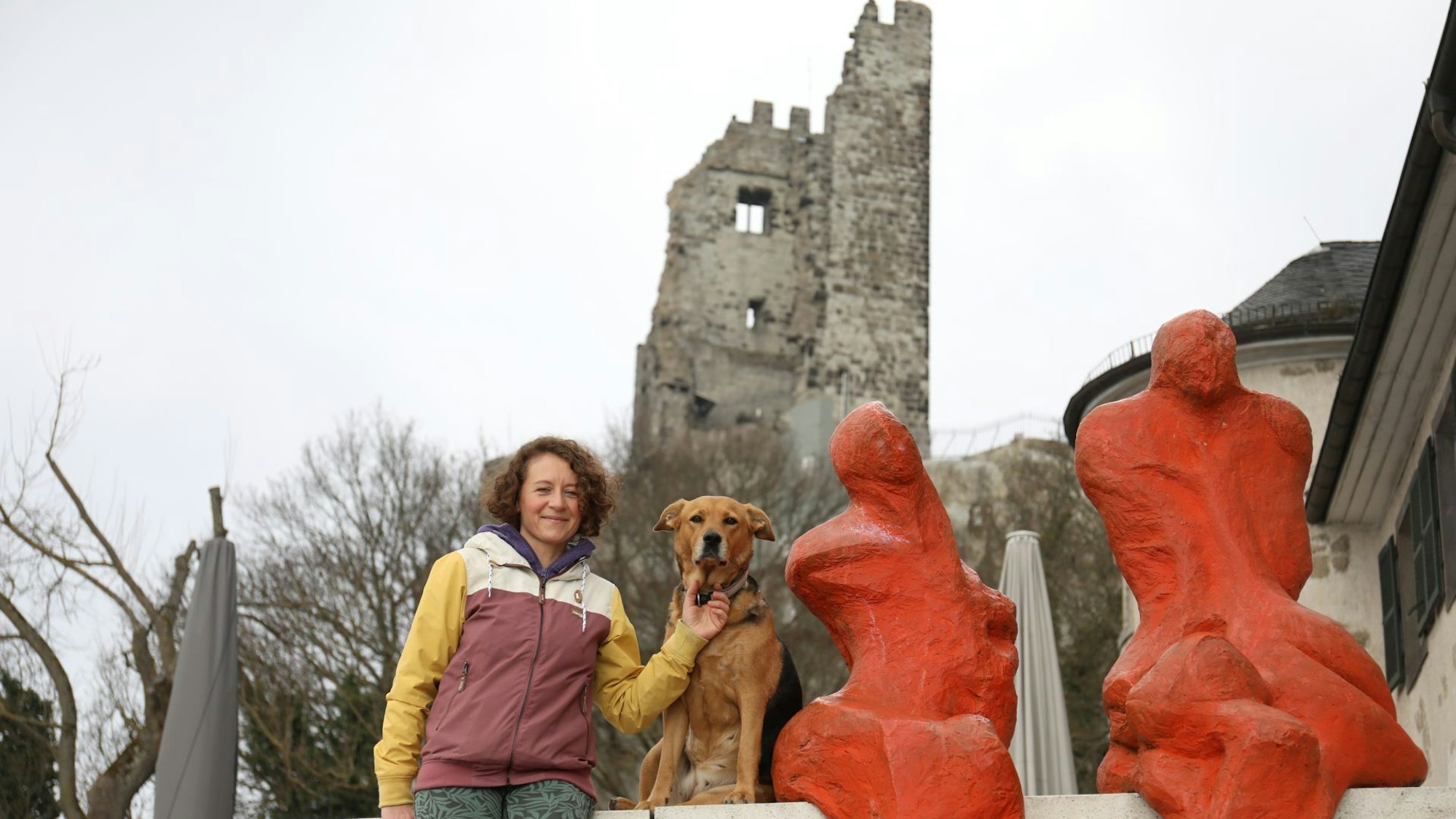 Eine Frau mit Hund neben zwei orange-roten Skulpturen, im Hintergrund die Ruine der Drachenburg.