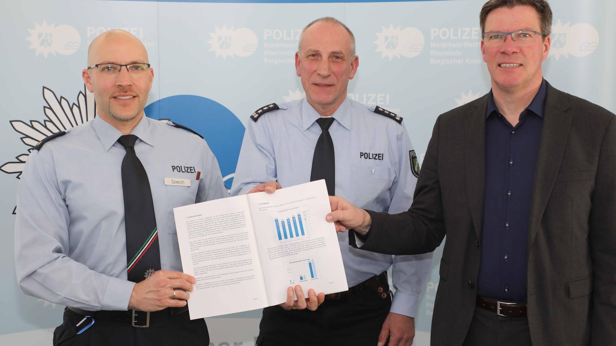 Markus Szech (Leiter Direktion Verkehr), Thomas Schliwitzki (Leiter Verkehrsdienst und Prävention & Opferschutz) und Landrat Stephan Santelmann halten die Verkehrsunfallbilanz für Rhein-Berg 2024.