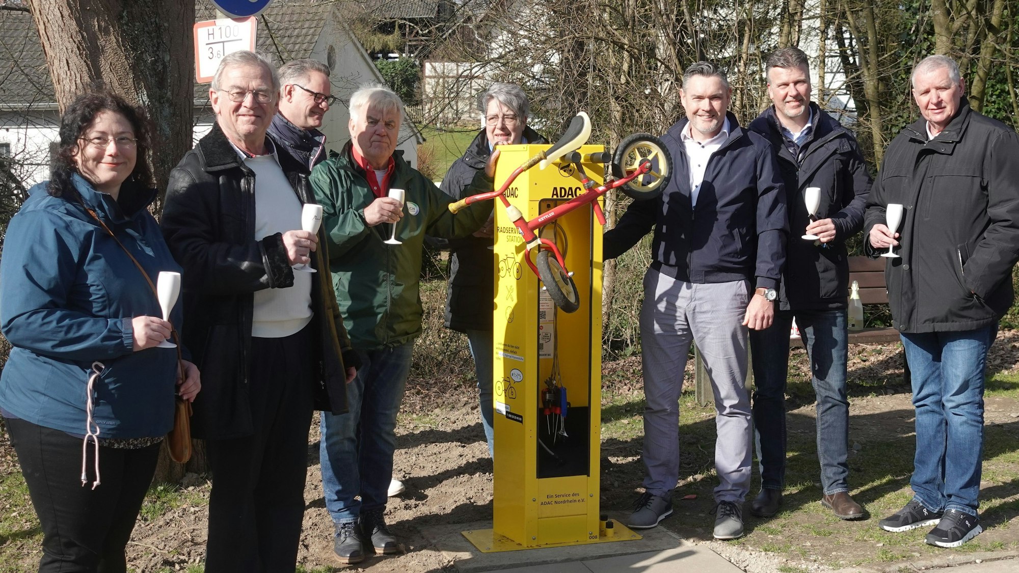 Acht Personen stehen links und rechts von einer gelben Säule mit Werkzeug und einem daran hängendem Kinder-Laufrad.