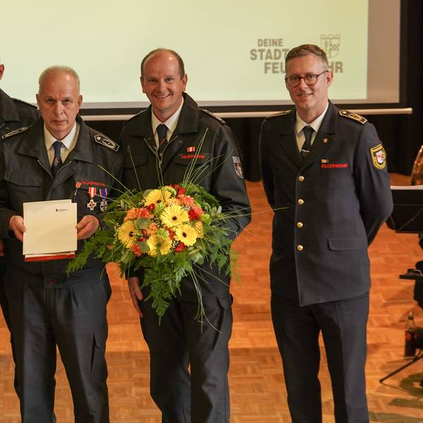 Gruppenbild mehrere Männer. Ein Feuerwehrmann hält eine Urkunde in der Hand. Ein weiterer Feuerwehrmann neben ihm einen Blumenstrauß.