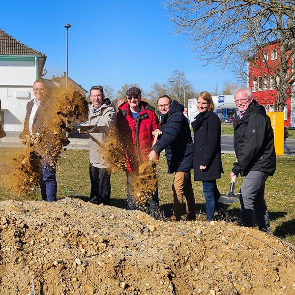 Zu sehen sind mehrere Männer und eine Frau, die mit Spaten an einer Baustelle Erde werfen.