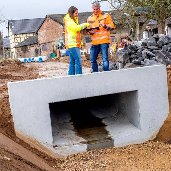 Eine Frau und ein Mann mit signalfarbenen Jacken stehen auf einem Rechteckprofil an einer Straßenbaustelle in Antweiler.