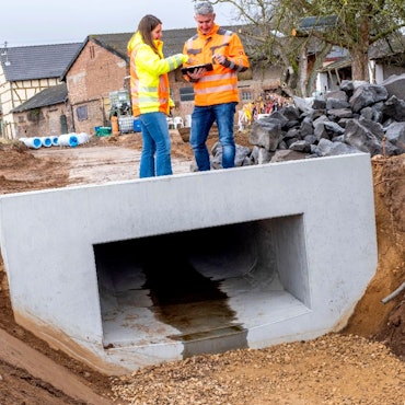 Eine Frau und ein Mann mit signalfarbenen Jacken stehen auf einem Rechteckprofil an einer Straßenbaustelle in Antweiler.