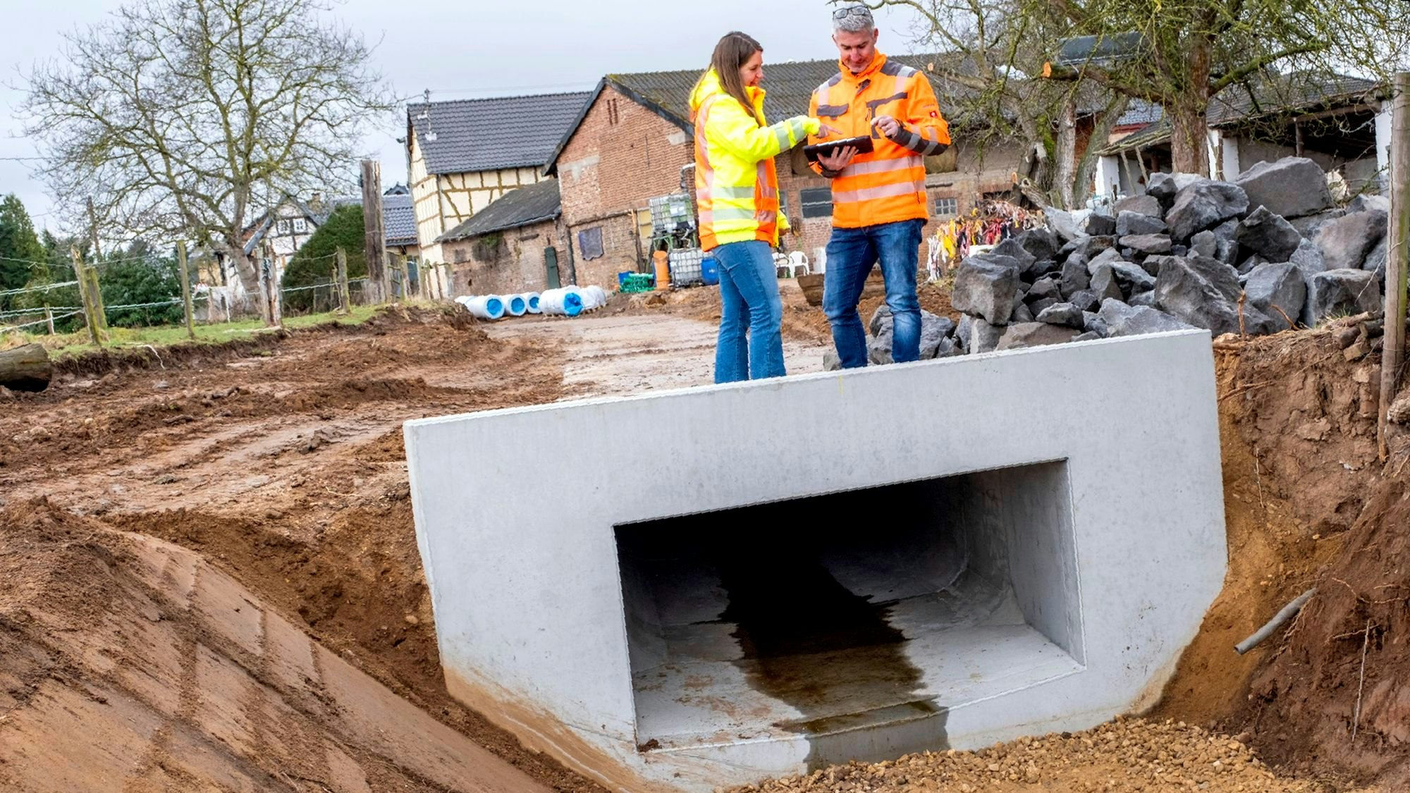 Eine Frau und ein Mann mit signalfarbenen Jacken stehen auf einem Rechteckprofil an einer Straßenbaustelle in Antweiler.