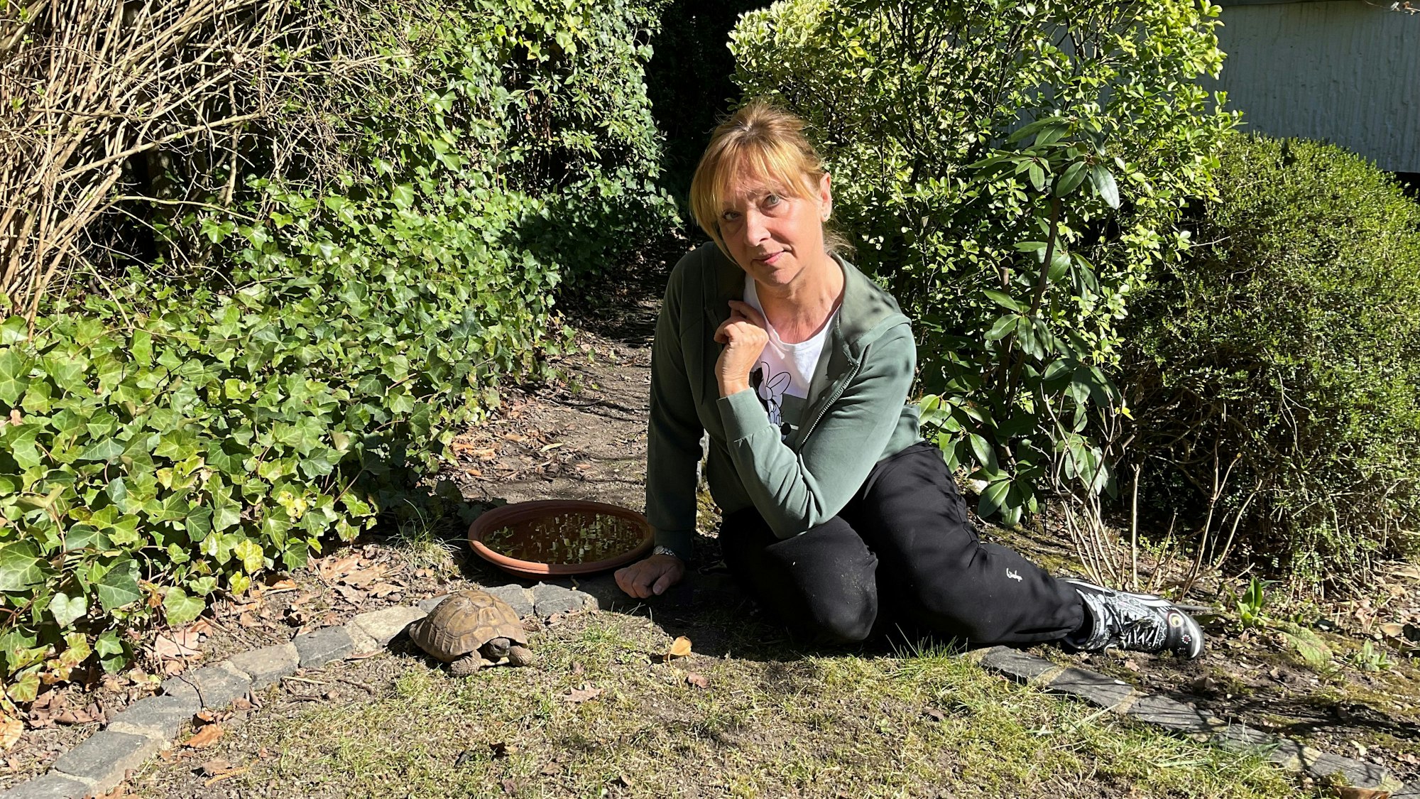 Eine Frau sitzt neben einer Landschildkröte in einem Garten.
