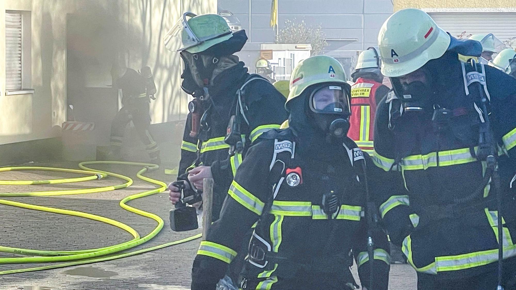 Das Bild zeigt die Euskirchener Feuerwehr im Einsatz. Im Hintergrund ist Rauch zu sehen, der aus einer geöffneten Tür ins Freie zieht.
