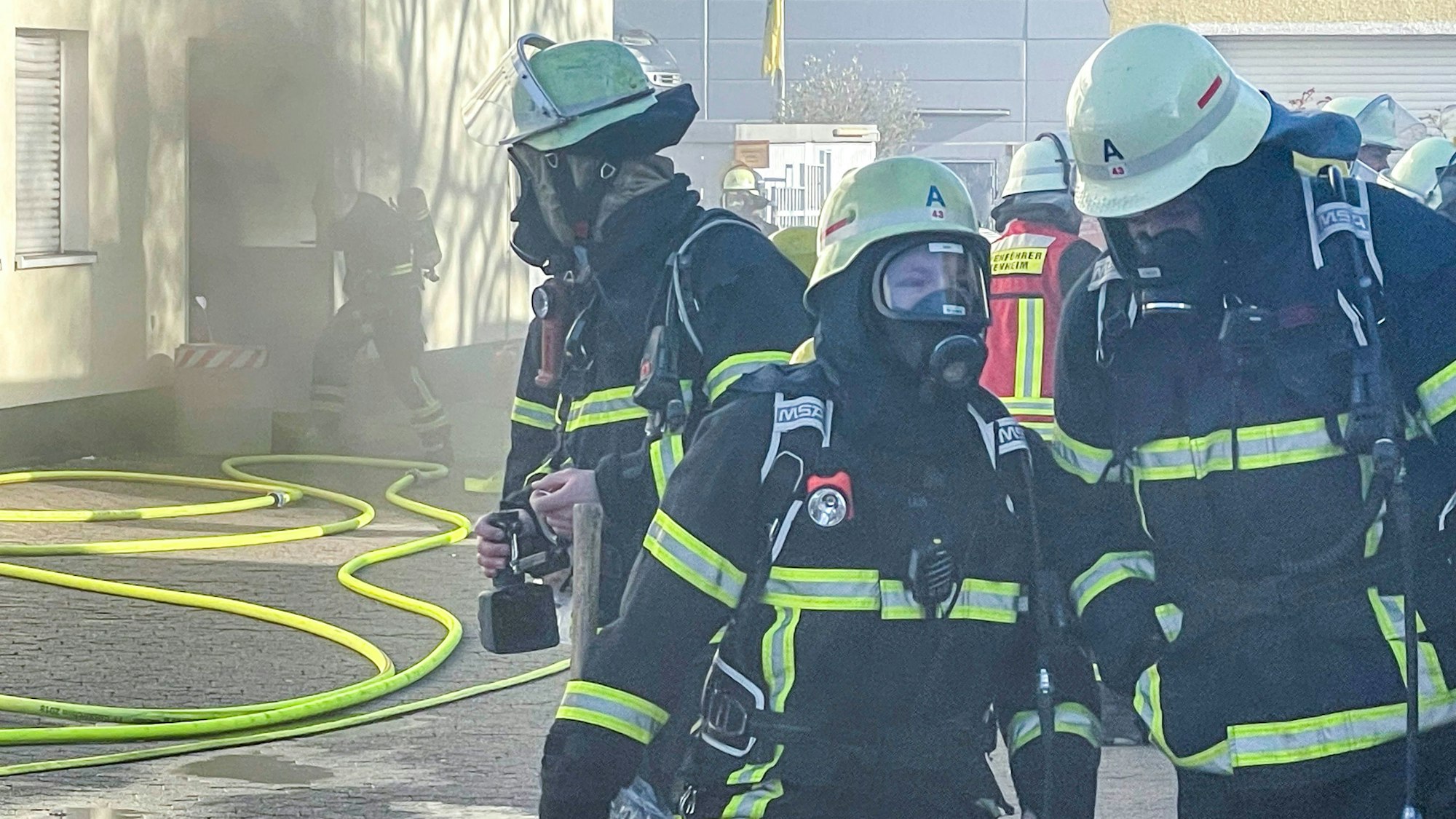 Das Bild zeigt die Euskirchener Feuerwehr im Einsatz. Im Hintergrund ist Rauch zu sehen, der aus einer geöffneten Tür ins Freie zieht.