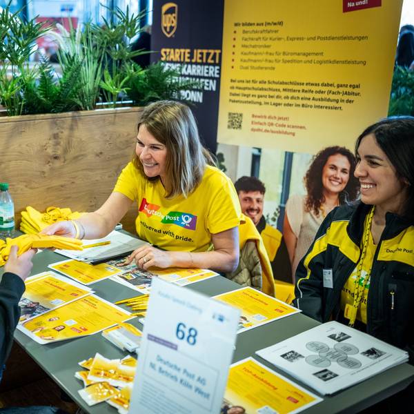 Ein Bewerber stellt dich der zwei Frauen von der Deutschen Post vor. Sie sitzen sich gegenüber an einem Tisch mit Werbeflyern.