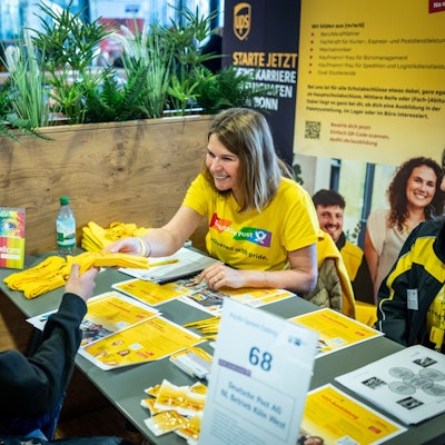 Ein Bewerber stellt dich der zwei Frauen von der Deutschen Post vor. Sie sitzen sich gegenüber an einem Tisch mit Werbeflyern.