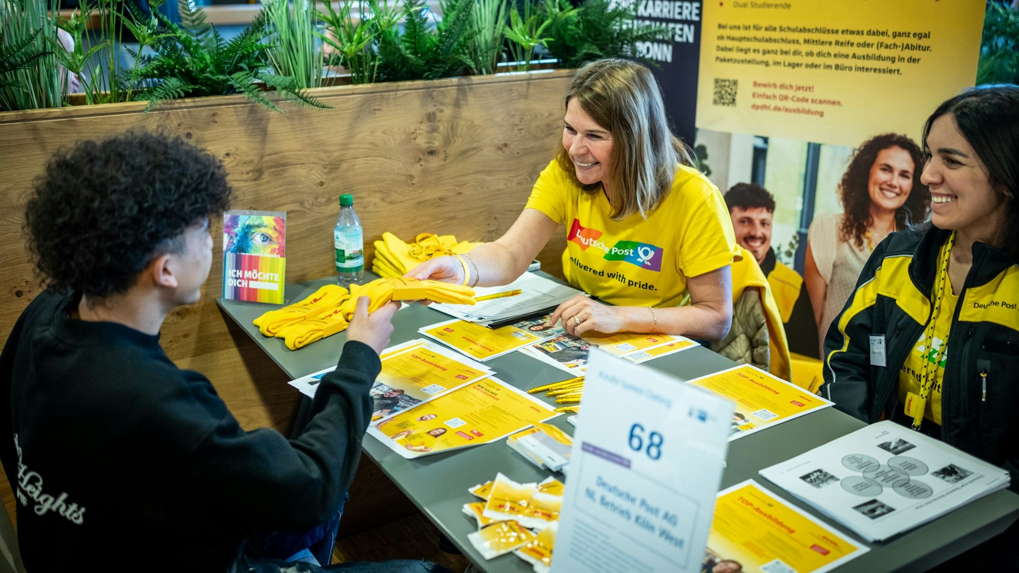 Ein Bewerber stellt dich der zwei Frauen von der Deutschen Post vor. Sie sitzen sich gegenüber an einem Tisch mit Werbeflyern.
