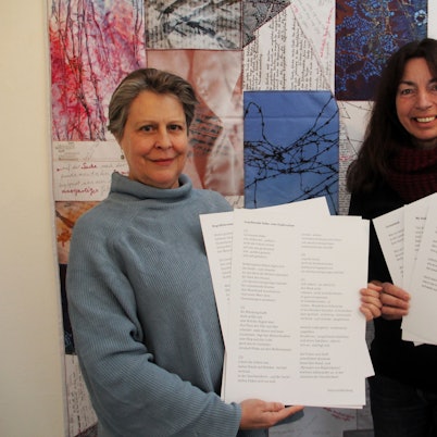 Zwei Frauen halten weiße Blätter mit Lyrik in den Händen. Im Hintergrund ist eine Patchwork-Decke zu sehen.