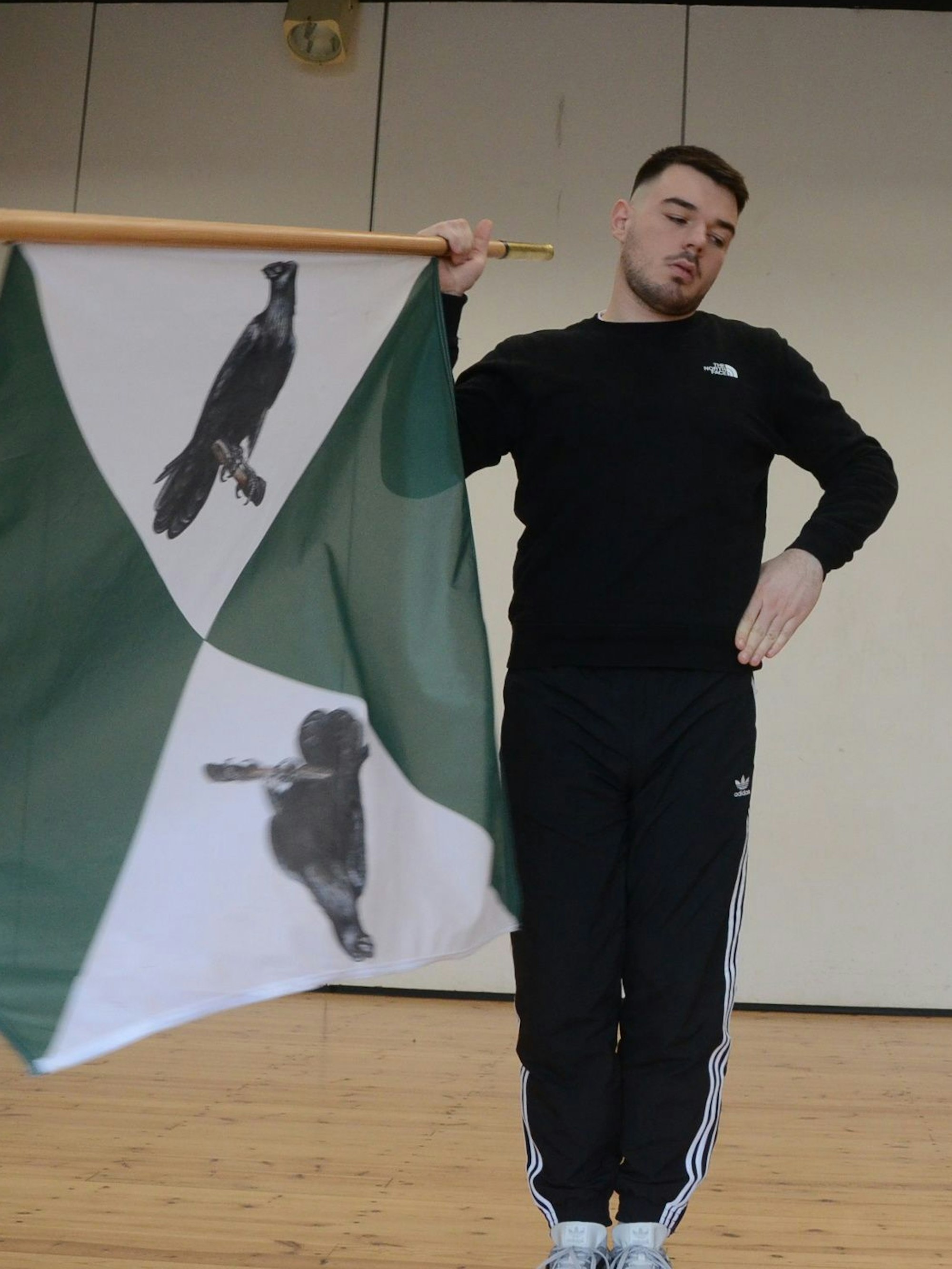 Ein junger Mann im schwarzen Trainingsanzug mit einer grün-weißen Fahne, auf die schwarze Krähen gedruckt sind.