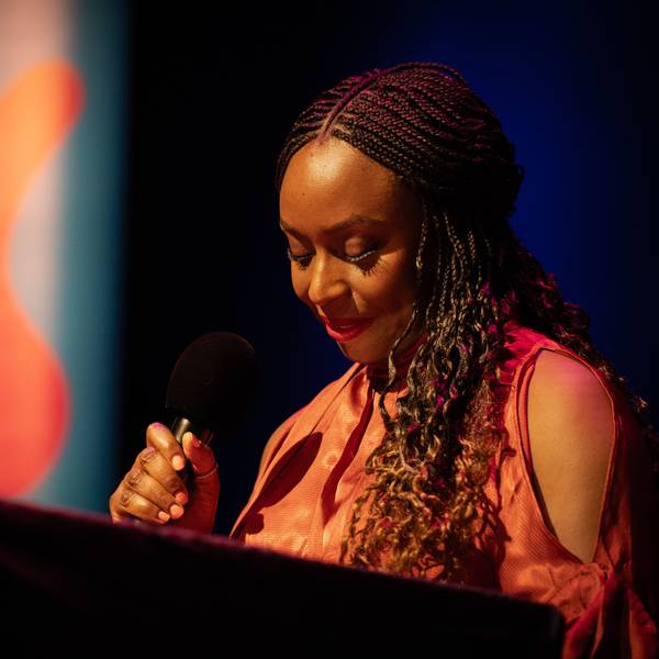 Chimamanda Ngozi Adichie spricht in der Stadthalle Mülheim in ein Mikrofon, sie hält die Augen gesenkt und trägt ein oranges Kleid.