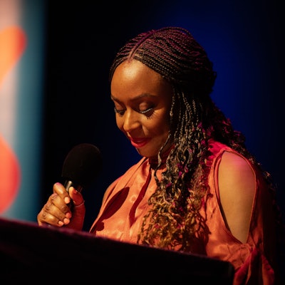 Chimamanda Ngozi Adichie spricht in der Stadthalle Mülheim in ein Mikrofon, sie hält die Augen gesenkt und trägt ein oranges Kleid.