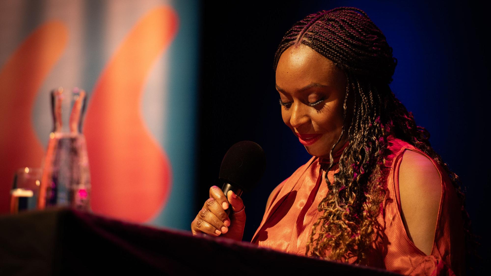Chimamanda Ngozi Adichie spricht in der Stadthalle Mülheim in ein Mikrofon, sie hält die Augen gesenkt und trägt ein oranges Kleid.
