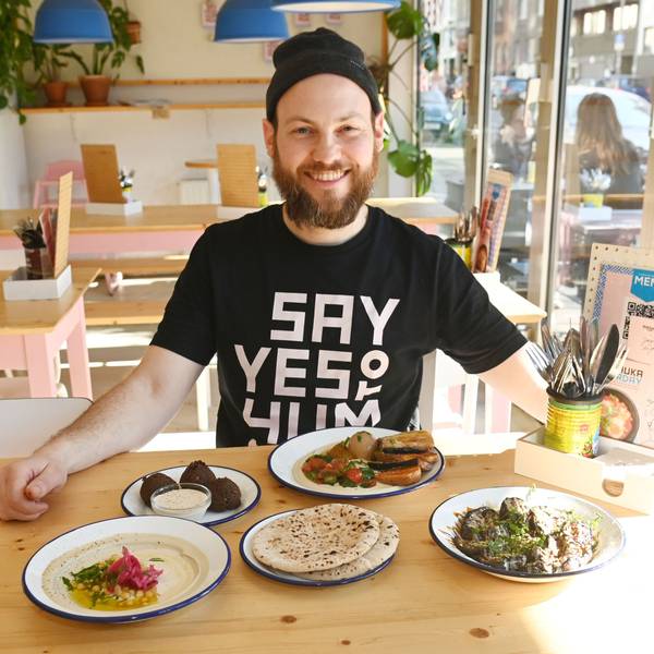 Wirtler ist mit einer Speisenauswahl an einem Tisch im Lokal zu sehen. Er trägt eine Strickmütze und schwarzes T-Shirt und lacht in die Kamera.