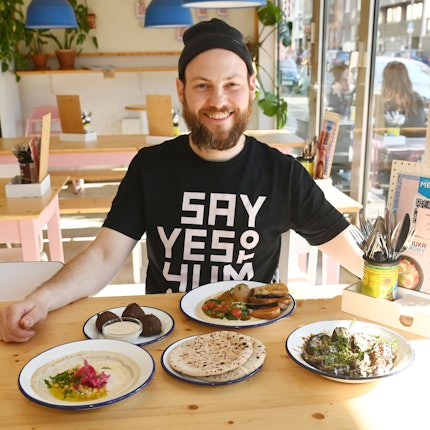 Wirtler ist mit einer Speisenauswahl an einem Tisch im Lokal zu sehen. Er trägt eine Strickmütze und schwarzes T-Shirt und lacht in die Kamera.