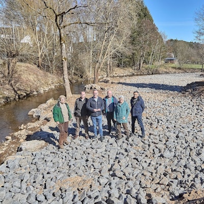 Sechs Personen stehen an einem Fluss auf einer Schotterfläche.