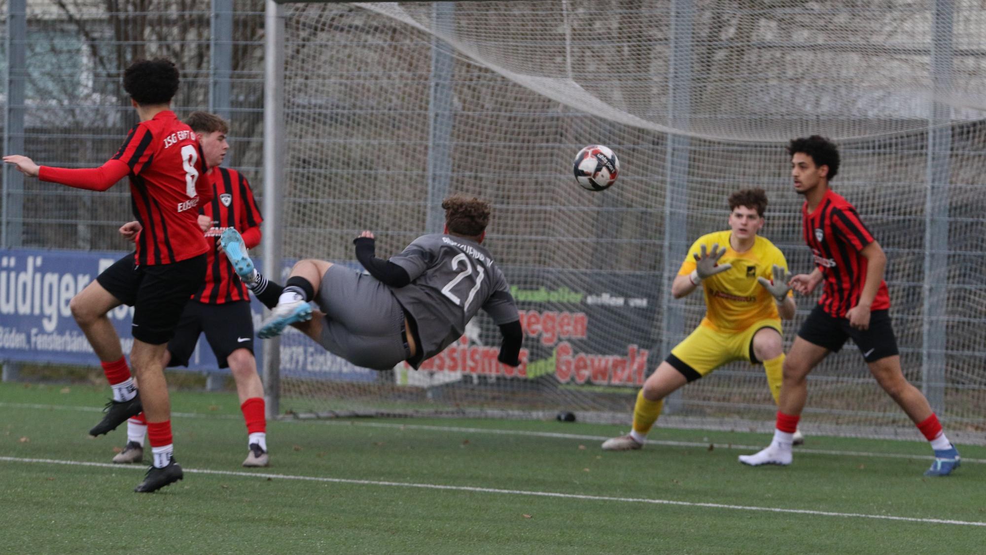 Ein Fußballspieler versucht per Fallrückzieher ein Tor zu erzielen. Den Ball wehrt ein Verteidiger noch vor dem Torwart ab.