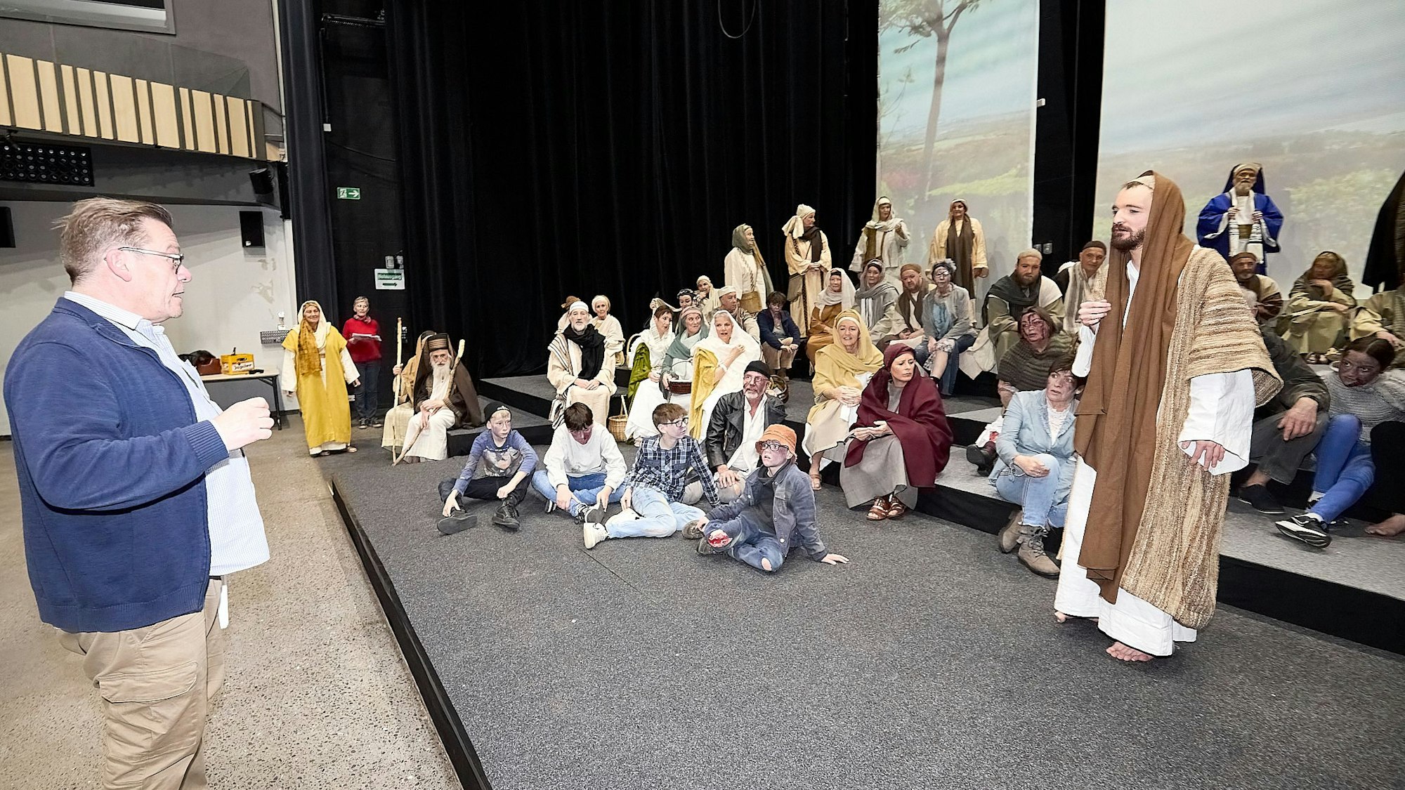 Der Regisseur spricht zu den Akteuren, die in ihren Kostümen auf den Treppenstufen der Bühne sitzen.
