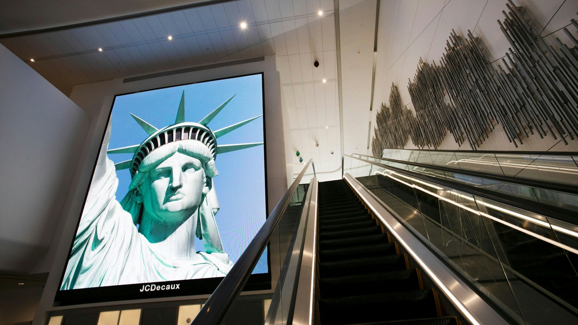 ARCHIV - 10.06.2020, USA, New York: Ein elektronisches Bildschirmbild der Freiheitsstatue begrüßt ankommende Reisende auf einer Rolltreppe im neuen Terminal B des Flughafens LaGuardia. (zu dpa: «Reisehinweise für USA jetzt mit Info zu Abschiebehaft») Foto: Mark Lennihan/AP/dpa +++ dpa-Bildfunk +++
