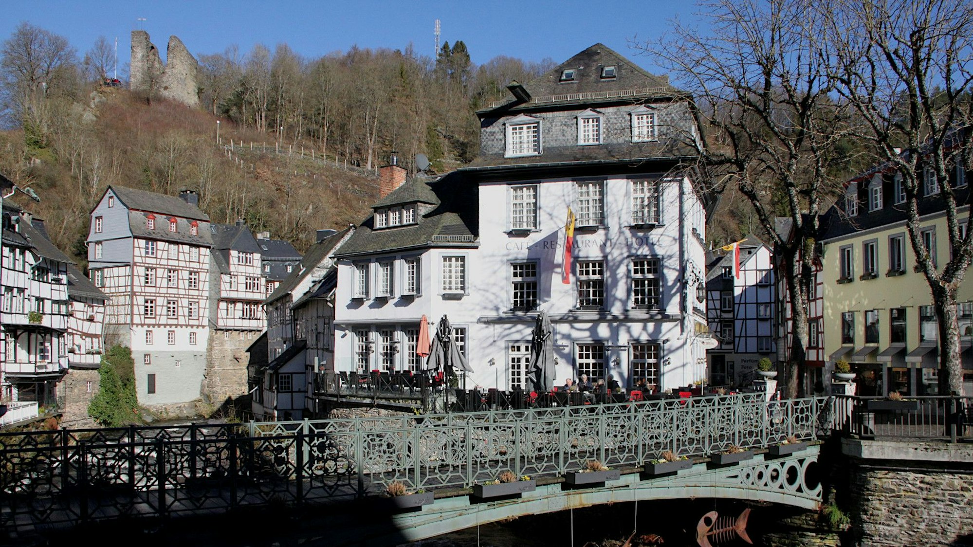 Das Bild zeigt Fachwerkhäuser und andere Gebäude in Monschau und im Hintergrund am Berghang die Ruine.