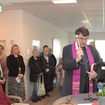 Das Bild zeigt den Priester mit dem Aspergill, die Gäste im Hintergrund schauen ihm zu.