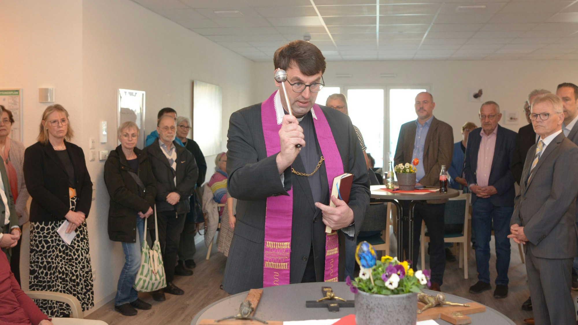 Das Bild zeigt den Priester mit dem Aspergill, die Gäste im Hintergrund schauen ihm zu.