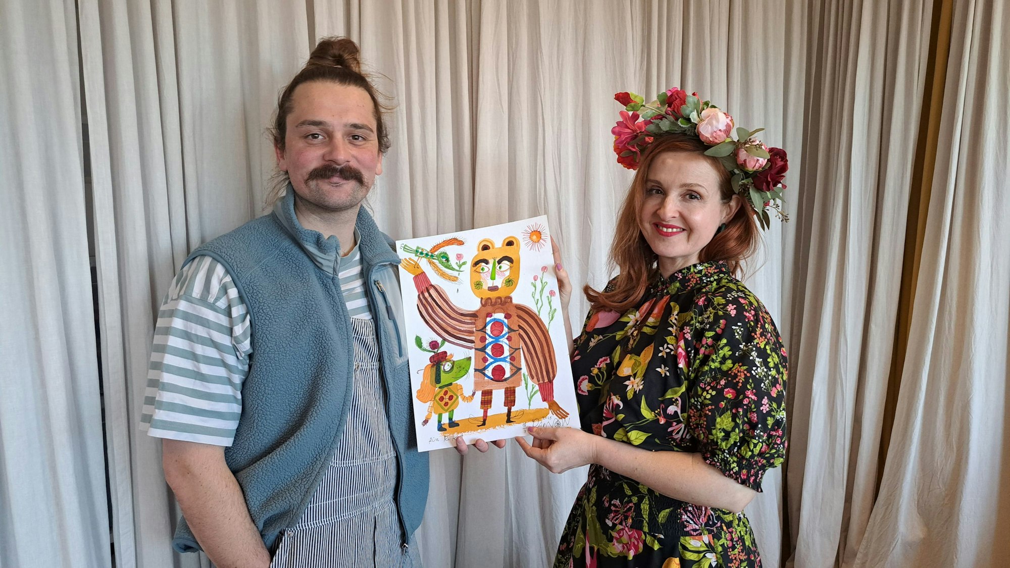 Ein Mann und eine Frau stehen vor einem beigen Vorhang. Sie halten ein gemaltes Bild zwischen sich.