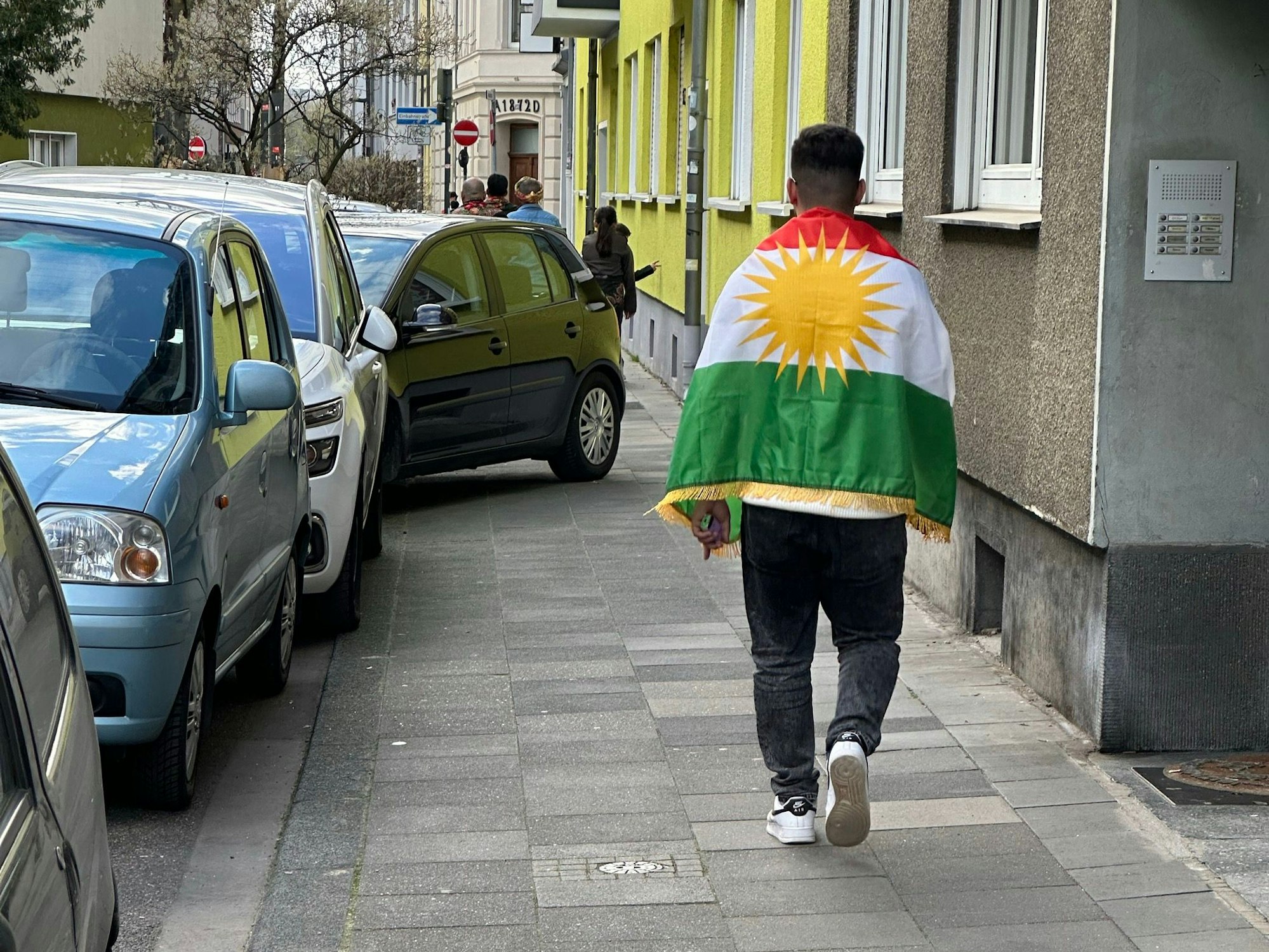 Ein mit einer kurdischen Fahne behangener Mann spaziert im Zuge des kurdischen Neujahrsfests durch Deutz, ein Auto im Hintergrund parkt halb auf dem Gehweg.