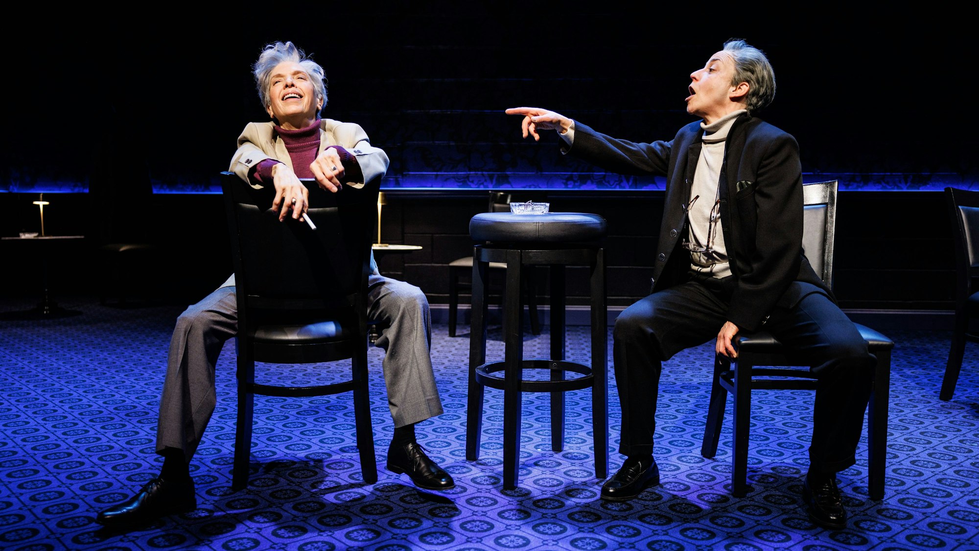 Helen Schneider (links) und Lucca Züchner sitzen auf Caféhausstühlen auf der Bühne der Stage 5 der New World Stages in New York. Schneider als Leonard Bernstein wirft lachend den Kopf zurück, Lucca Züchner als Herbert von Karajan, gestikuliert erregt mit dem Zeigefinger.