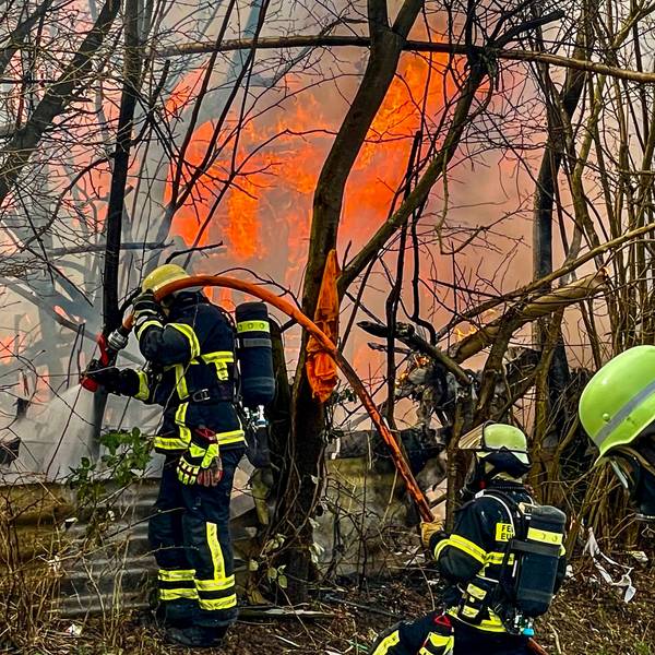Das Bild zeigt drei Feuerwehrleute, die den Brand bekämpfen. Im Hintergrund sind Flammen zu sehen.