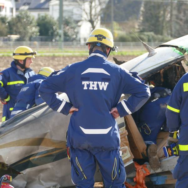 Männer in Overalls des THW stehen vor dem Wrack des abgestürzten Flugzeuges am Flugplatz in Sankt Augustin.