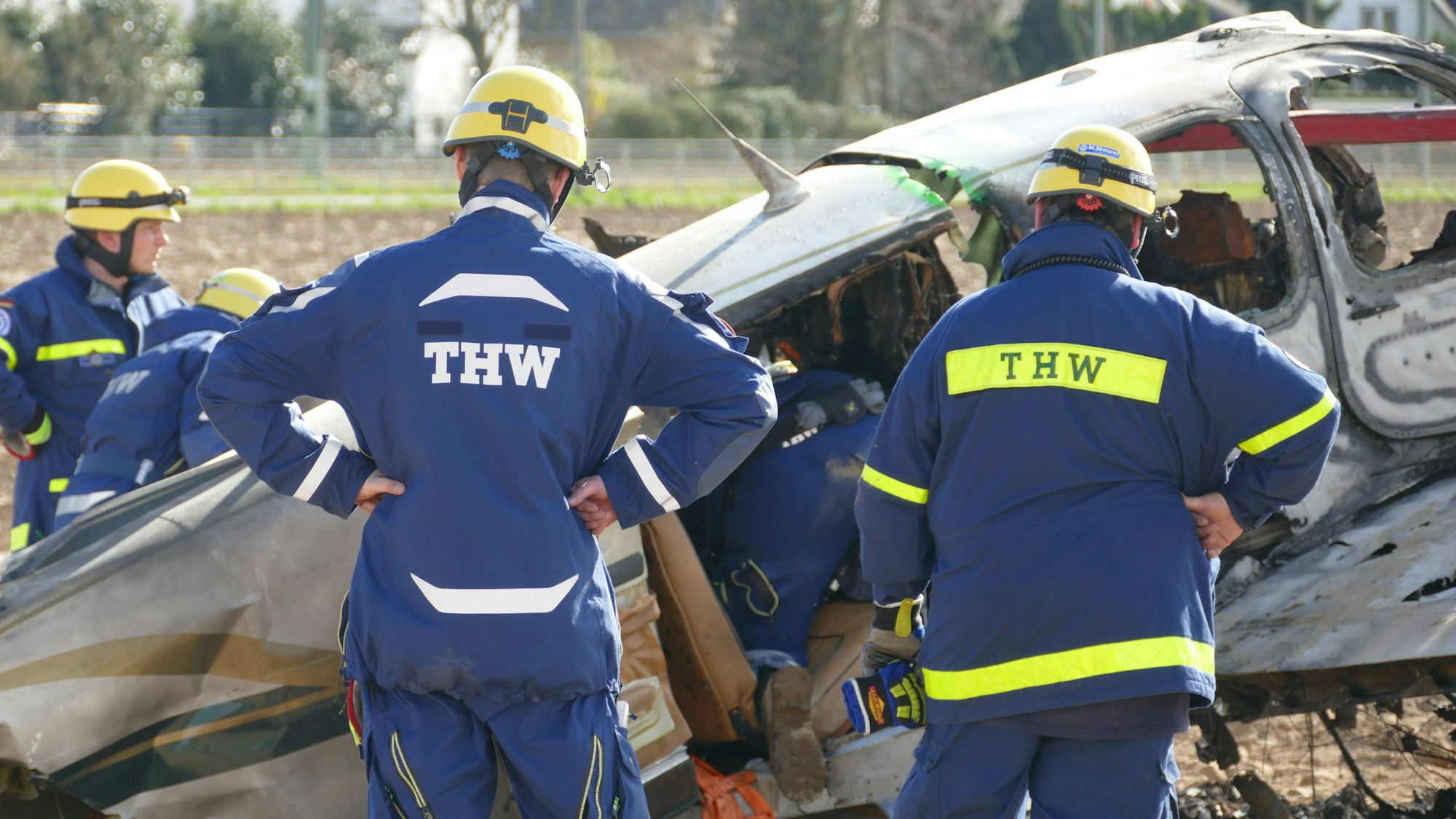 Männer in Overalls des THW stehen vor dem Wrack des abgestürzten Flugzeuges am Flugplatz in Sankt Augustin.