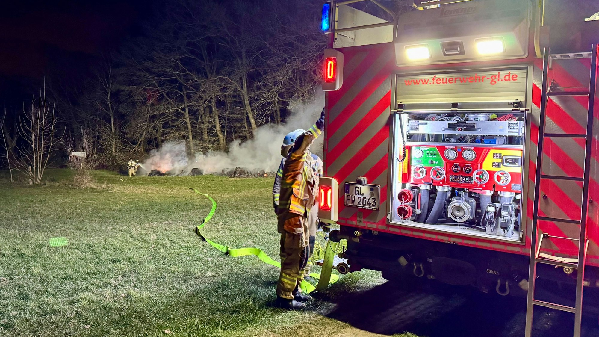 Ein Feuerwehrfahrzeug steht auf einer Wiese, am Waldrand dahinter sind Feuerwehrleute und Qualm zu sehen.