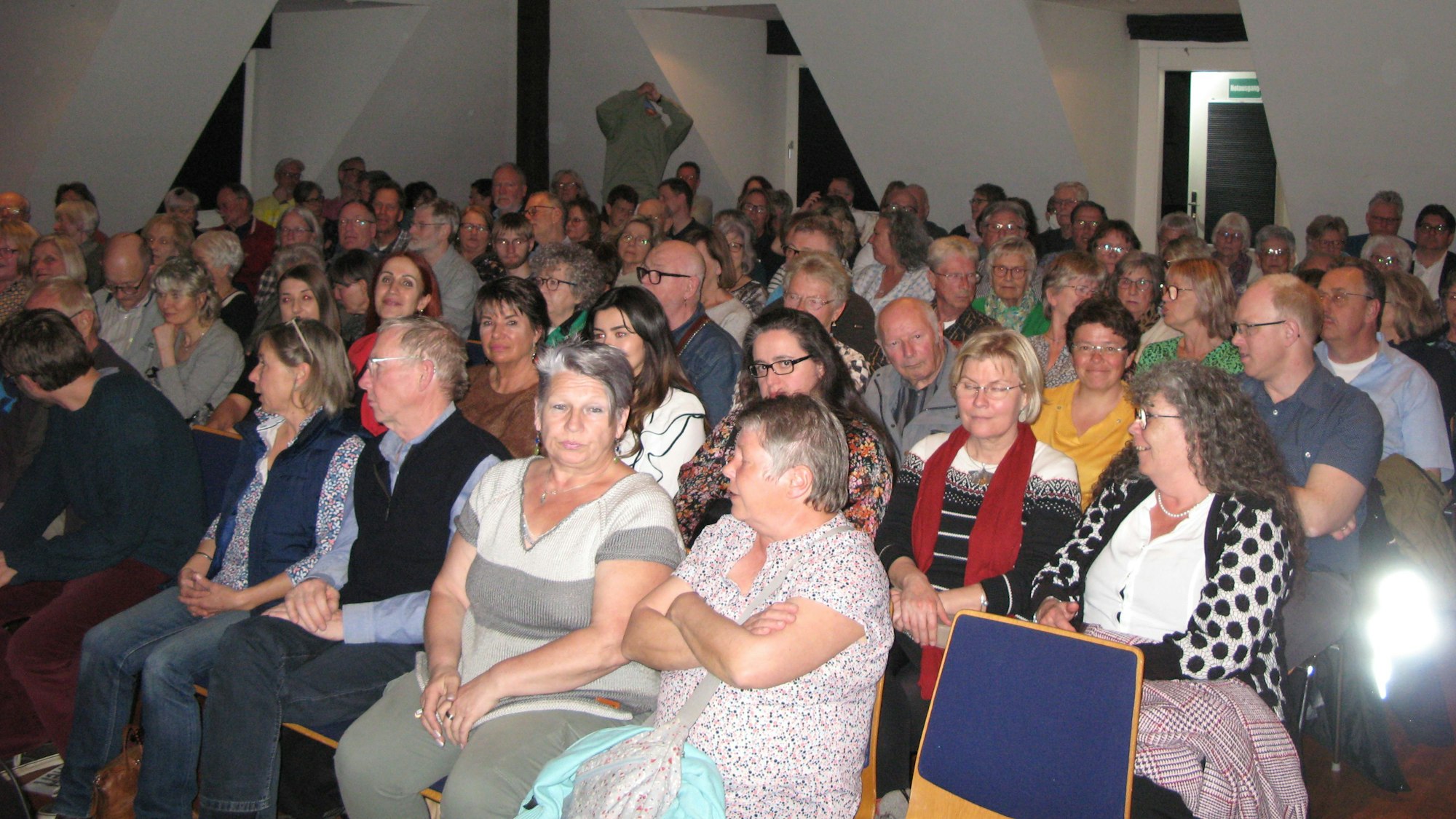 Sieben besetzte Stuhlreichen unter einer Dachstuhldecke.