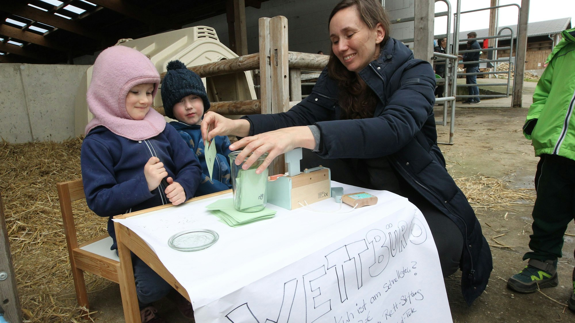 Zwei kleine Kinder hocken mit ihrer Mutter an einem kleinen Tisch mit einem Plakat, auf dem „Wettbüro“ steht.
