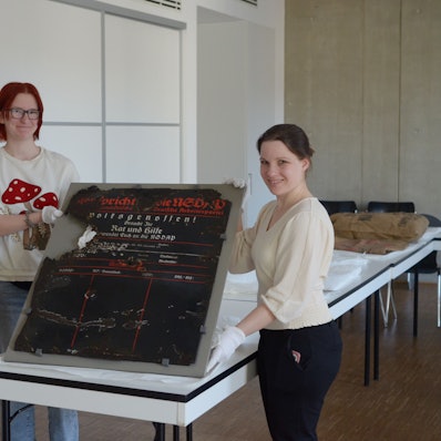 Das Foto zeigt die drei Frauen. Zwei von ihnen halten eine historische Infotafel.