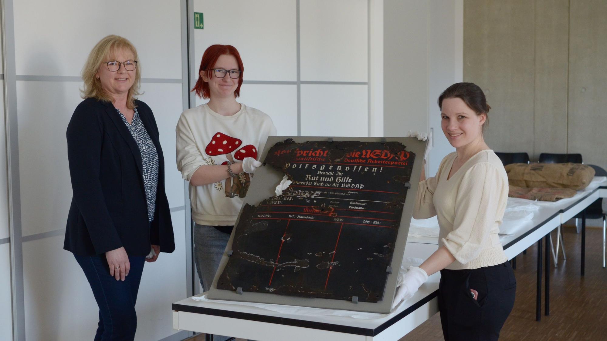 Das Foto zeigt die drei Frauen. Zwei von ihnen halten eine historische Infotafel.