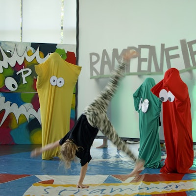 Ein Kind schlägt auf einer Bühne ein Rad. Im Hintergrund steht auf einer Leinwand das Wort „Rampenfieber“.