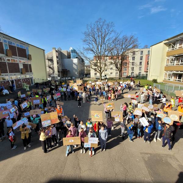 Zahlreiche Schulkinder stehen auf einem Schulhof und halten selbsterstellte Plakate in die Höhe.