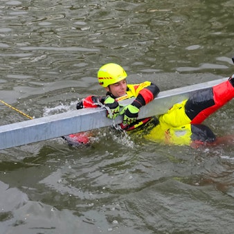 Zu sehen ist ein Strömungsretter der DLRG, der sich in einem Gewässer an einen Metallbalken klammert.