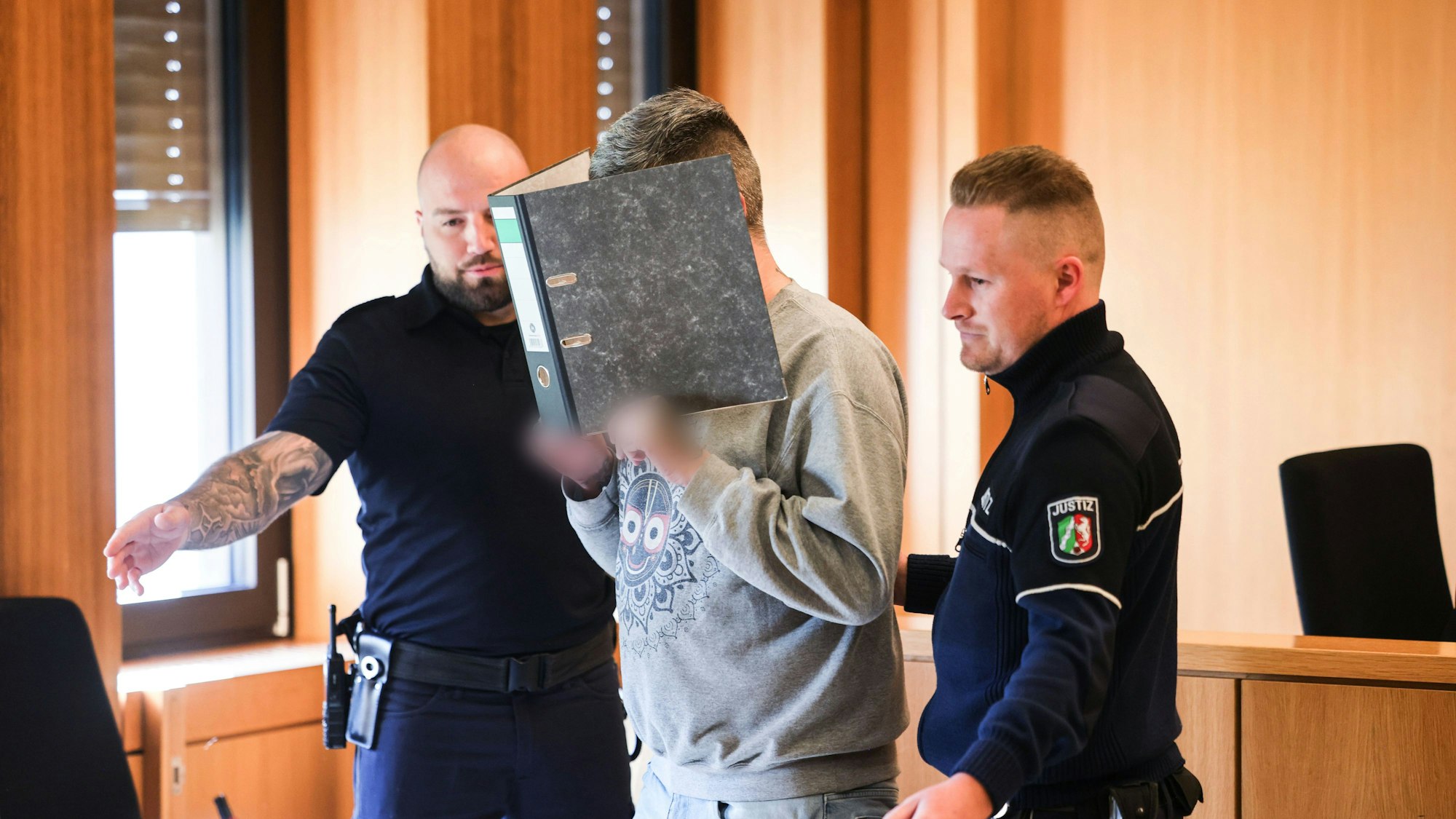 Der angeklagte Krankenpfleger hält sich einen Aktenordner vor sein Gesicht, während er von Justizangestellten zu seinem Platz gebracht wird.