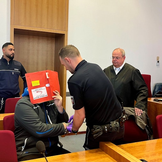 Das Foto zeigt den Angeklagten, der sein Gesicht mit einer Kladde verdeckt. Ein Justizwachtmeister löst ihm die Handfesseln, der Verteidiger beobachtet die Szene.