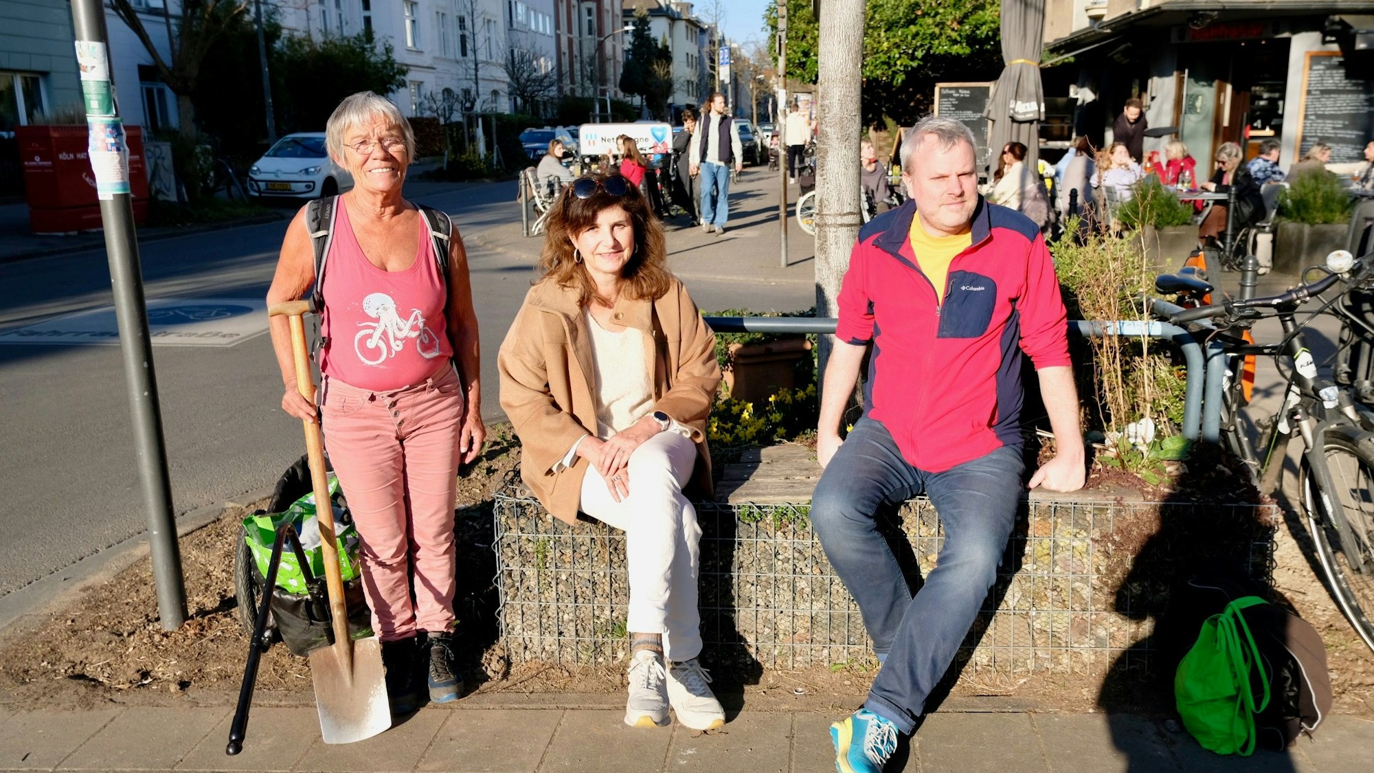 Zwei Frauen und ein Mann sitzen im Sonnensschein auf einem etwas hochgelegenen Beet.