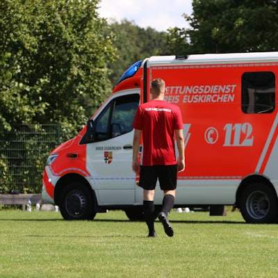 Ein Fußballer trottet in Richtung eines Rettungswagens, der auf dem Rasen eines Sportplatzes steht.