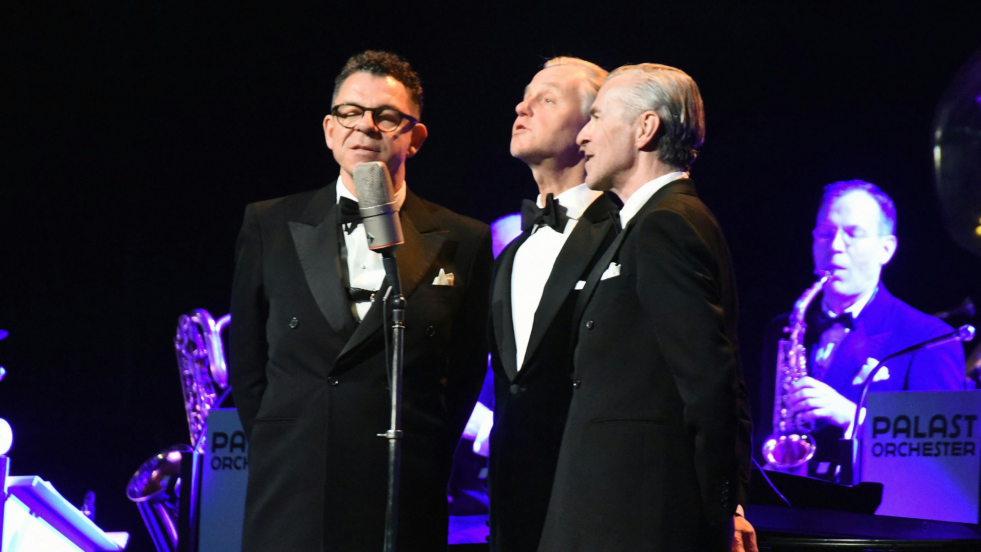 Sänger Max Raabe mit zwei Musikern des Palastorchesters auf einer Bühne.