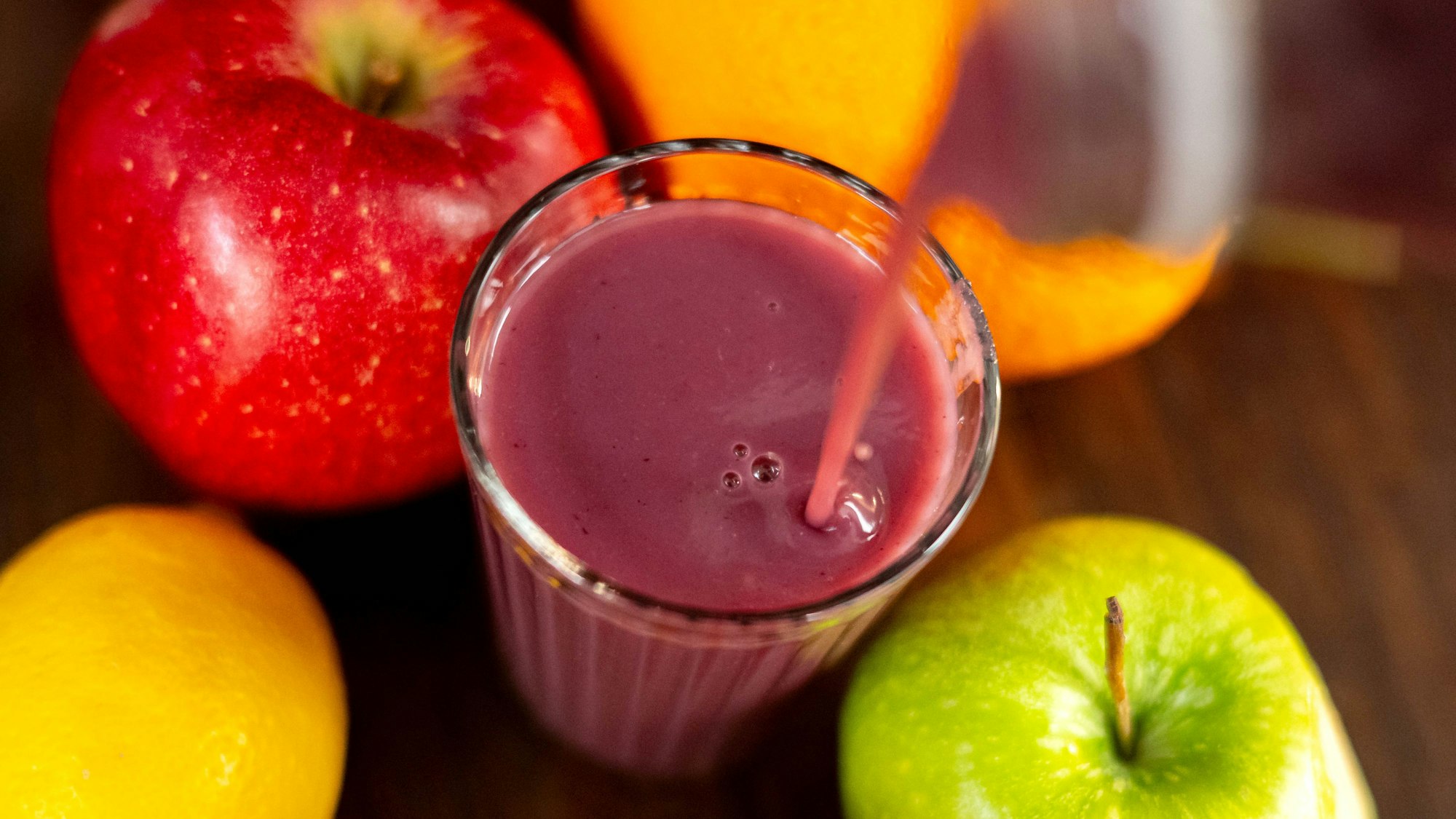 In ein Glas wird ein Smoothie eingegossen. Das Glas steht auf einem Holztisch inmitten von Obst. (Symbolbild)